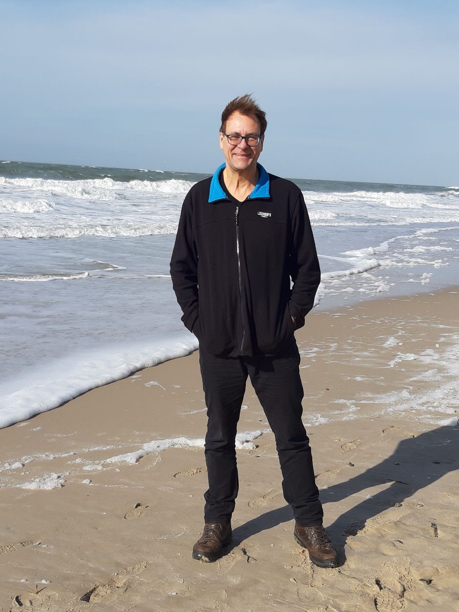 Andreas Dernehl steht am Strand, hinter ihm die Brandung